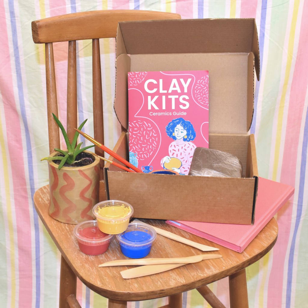 An open box sits on a wooden stool, set against a pastel striped background. Inside the box, there is a lump of clay and some paintbrushes sticking out, beside a leaflet that reads 'Clay Kits, Ceramics Guide'. Around the box are a notebook, wooden clay tools, three plastic pots with colourful contents and a small plantpot with a plant inside.